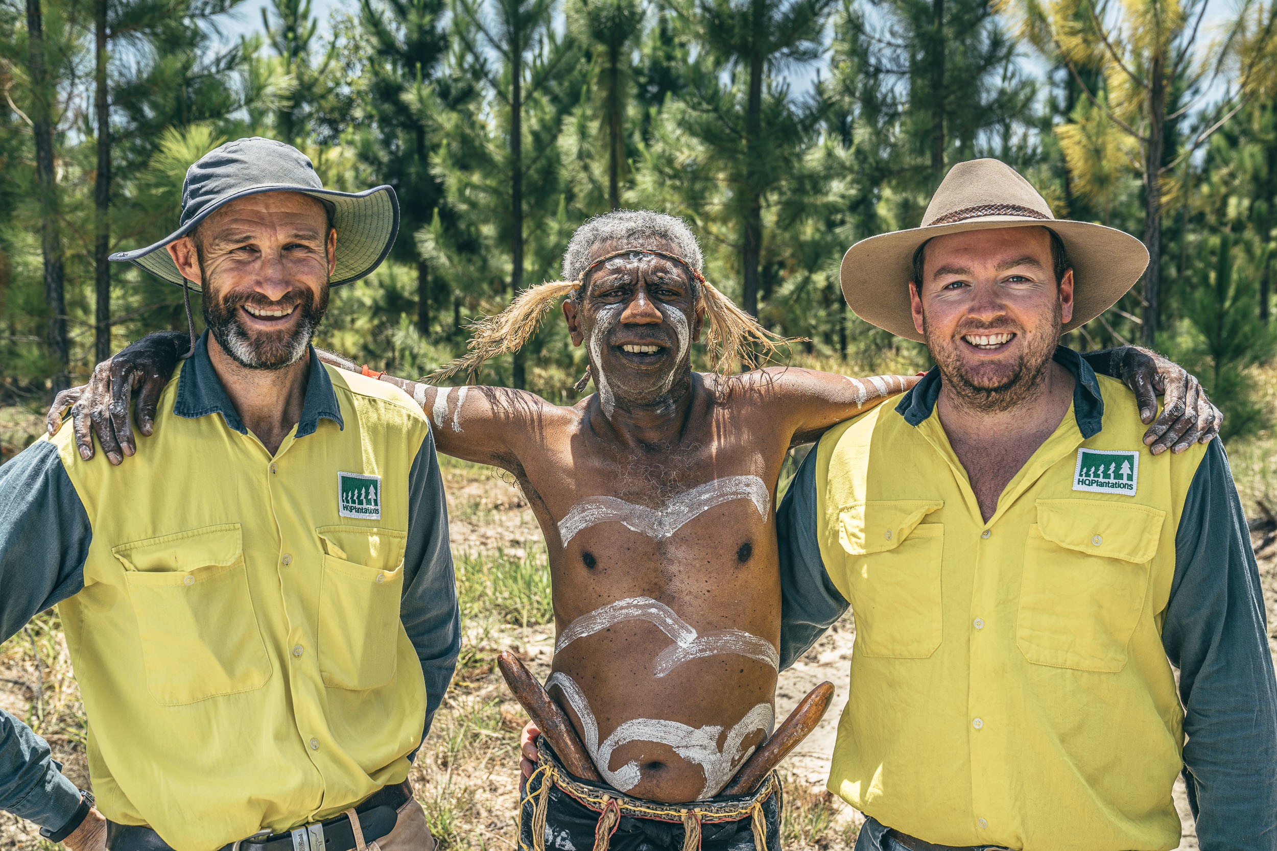 Group photo of a smiling Butchulla man in ceremonial dress and body painting with two smiling employees
